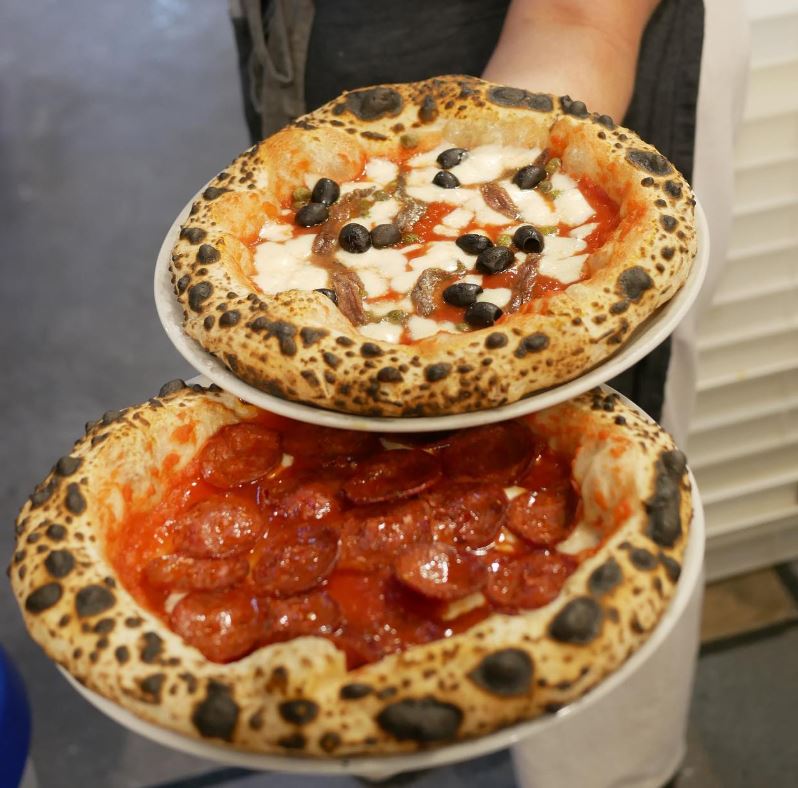 Person holding a pepperoni pizza and a pizaa with olives and anchovies